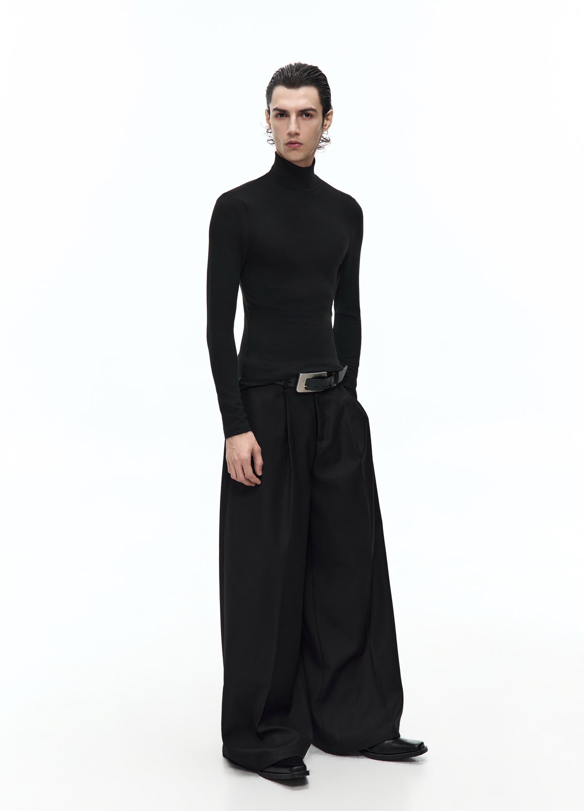 Front view of a model in a fitted black high-neck top with long sleeves, worn with a belt and very wide black trousers.