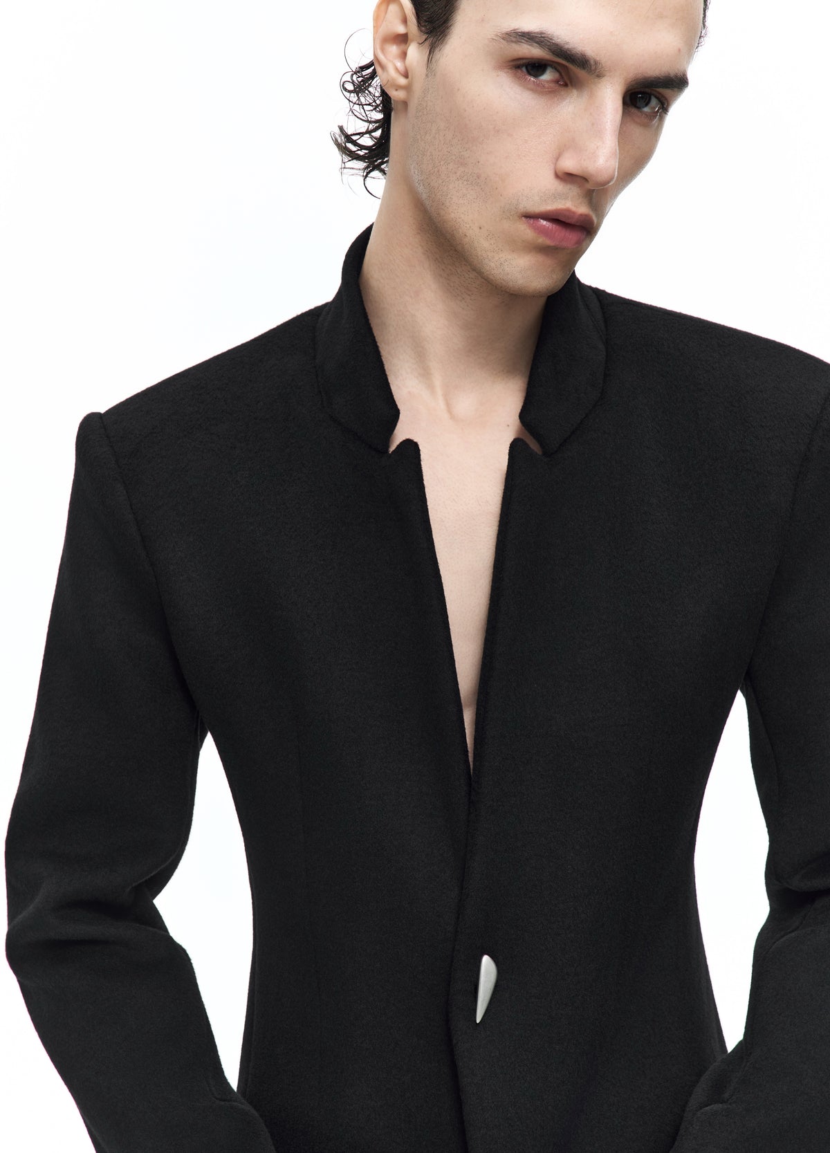 Close front view of the black jacket, showing the shaped collar, deep opening and small curved silver-tone fastening at the waist.
