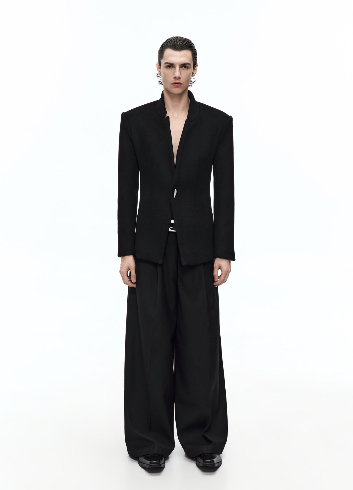 Front view of a model in a fitted black jacket with a high notched collar and small silver-tone front fastening, worn with wide trousers.