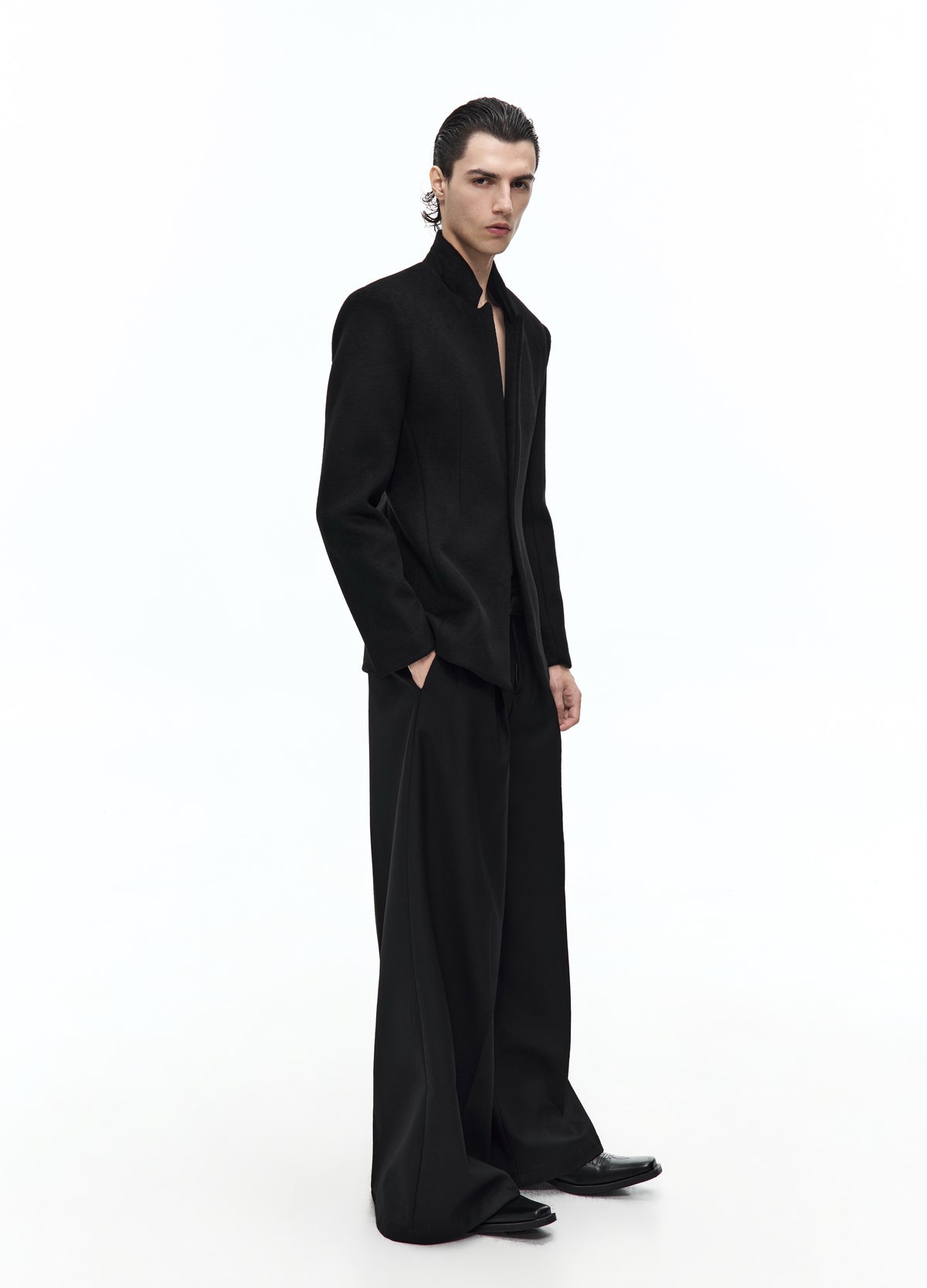 Side view of the fitted black high-collar jacket, showing the narrow waist and hem at the upper thighs above wide black trousers.