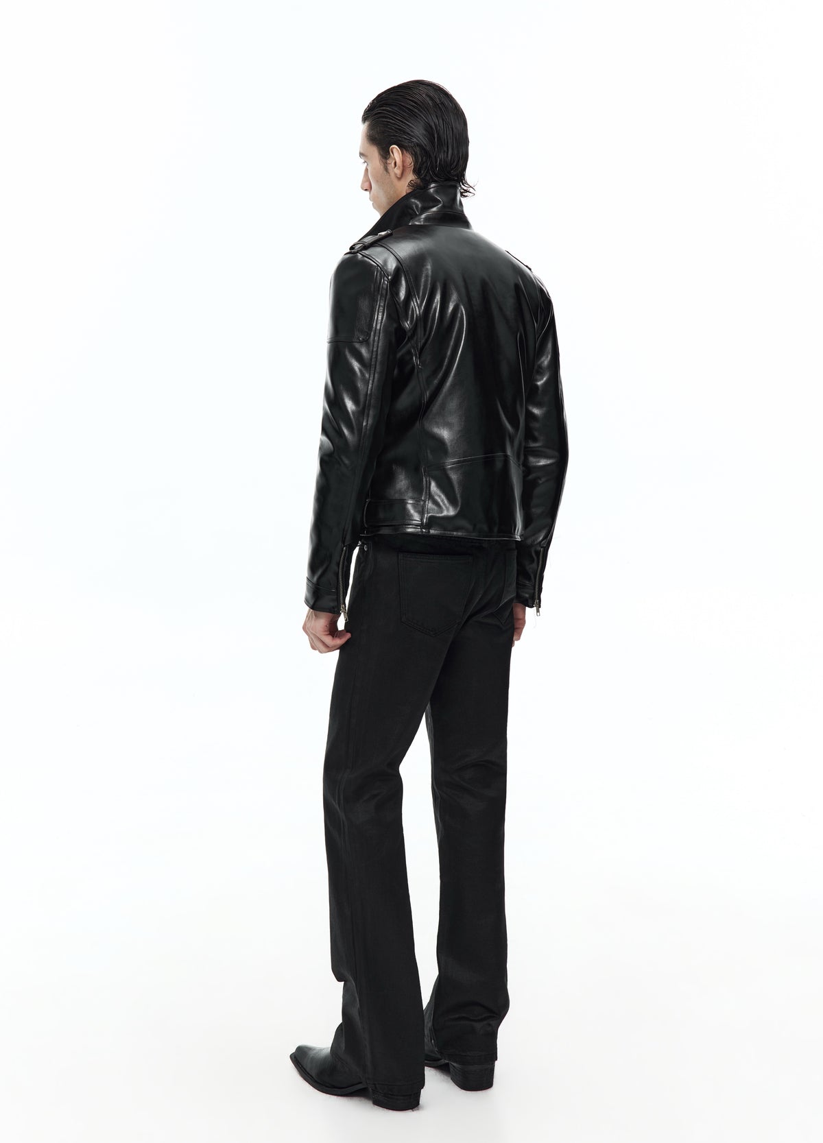 Rear three-quarter view of the black biker jacket, showing the back panels, long sleeves and hem above black trousers.