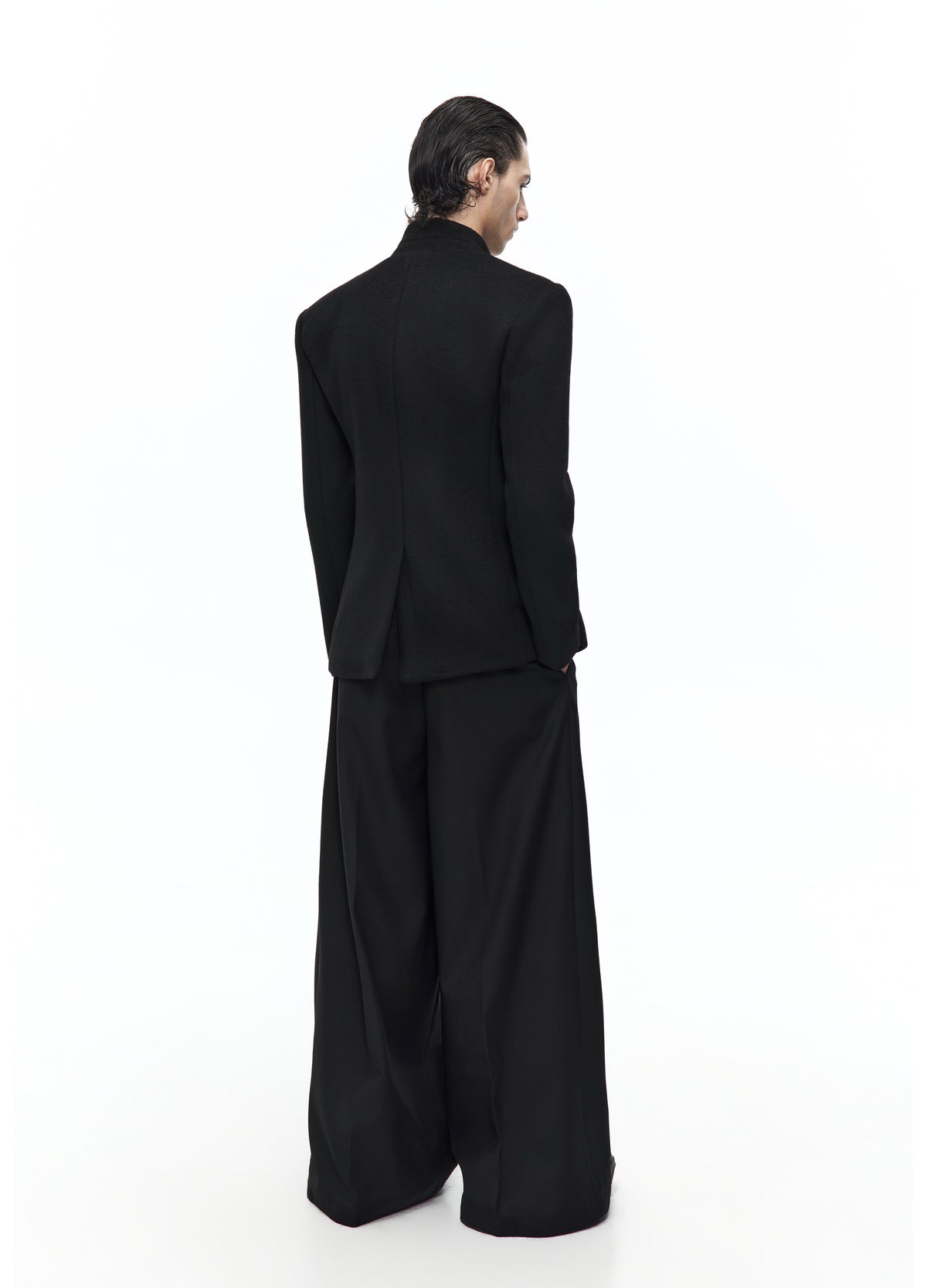 Rear three-quarter view of the black high-collar jacket, showing the shaped waist and rear vent above wide black trousers.